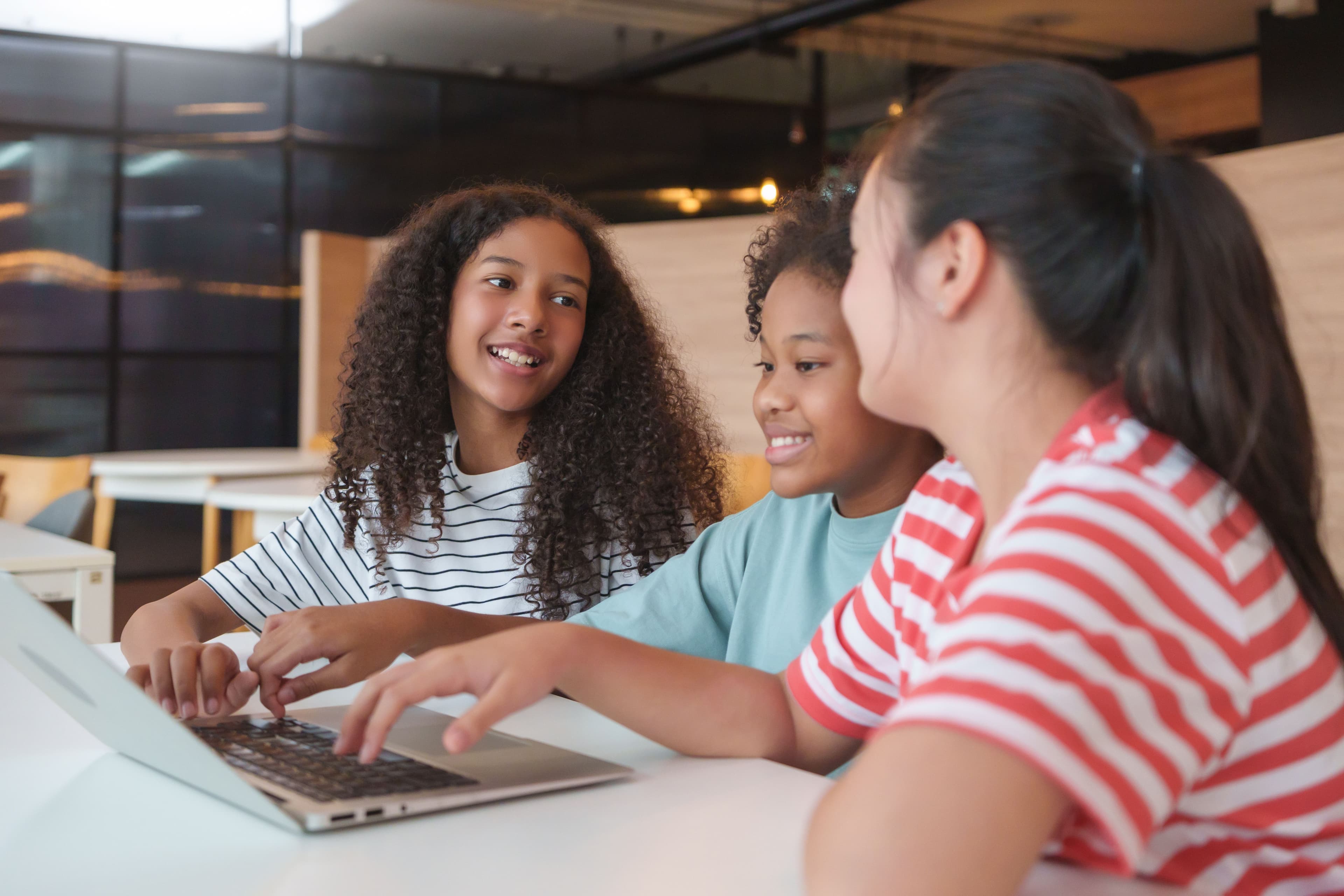 Three smiling children coding together on a laptop at a bright workshop table.