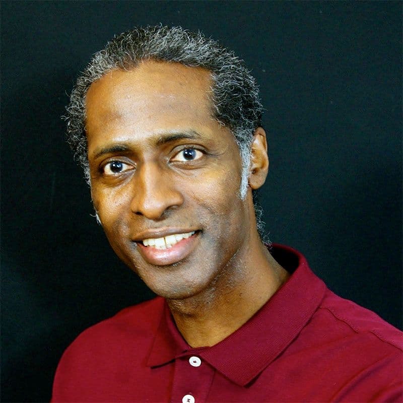Portrait of John Henry Thompson, short greying curls, dark backdrop, wearing a maroon polo.